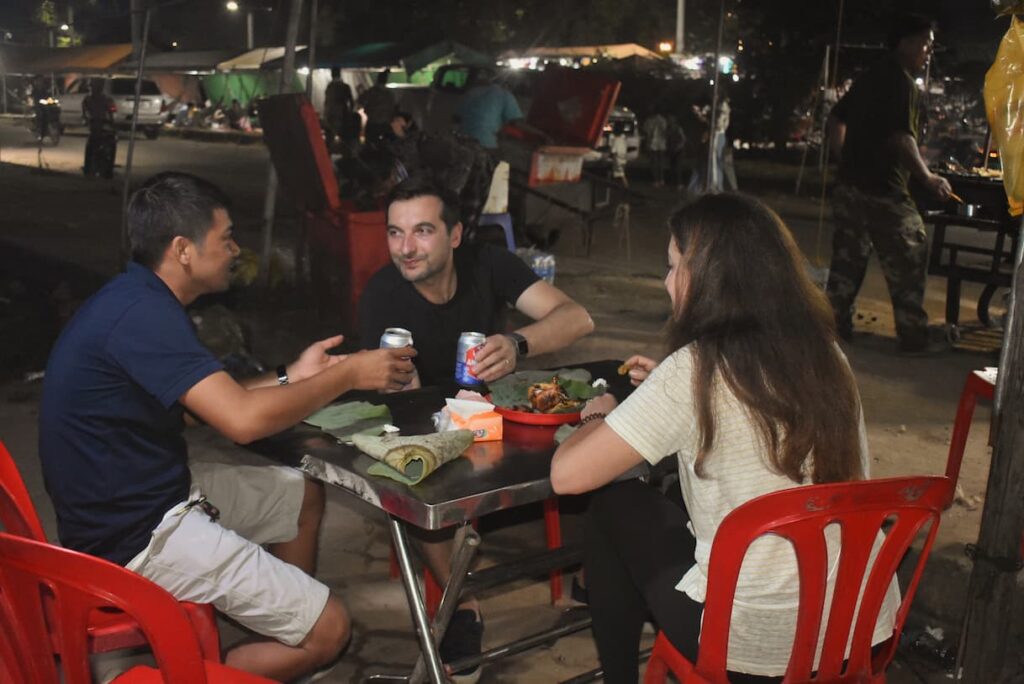 Guide and guests eating grilled food at a metal table in the Siem Reap night market